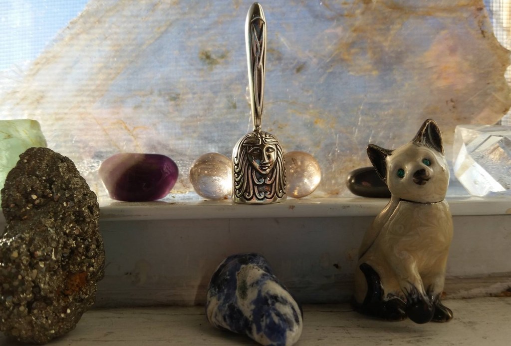 Product photo of a tiny silver bell engraved with a goddess face and trinity knot, backlit against a window with radiant stones surrounding it.